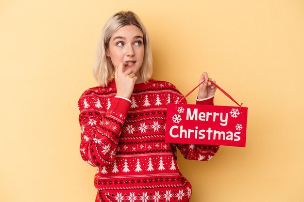 Junge kaukasische Frau, die ein Plakat der frohen Weihnachten auf gelbem Hintergrund isoliert hält, entspannte sich beim Nachdenken über etwas, das einen Kopienraum betrachtet.