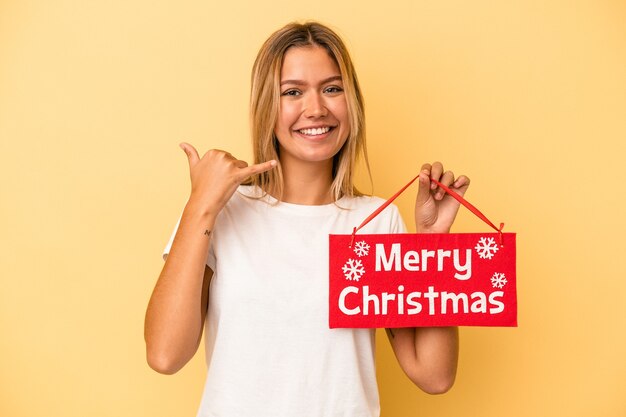 Junge kaukasische Frau, die ein Plakat der frohen Weihnachten hält, das auf gelbem Hintergrund lokalisiert wird, das eine Handyanrufgeste mit den Fingern zeigt.