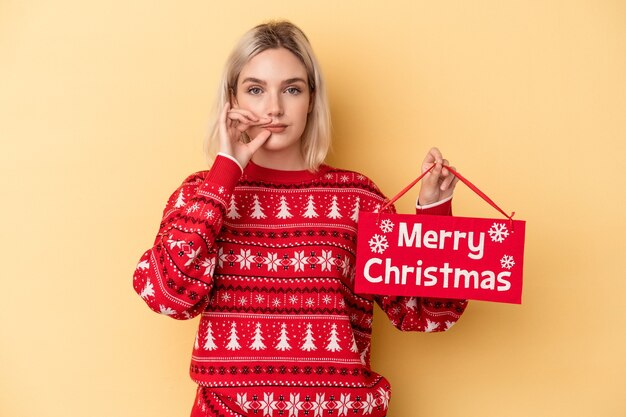 Junge kaukasische Frau, die ein Plakat der frohen Weihnachten hält, das auf gelbem Hintergrund mit Fingern auf den Lippen lokalisiert wird, die ein Geheimnis halten.