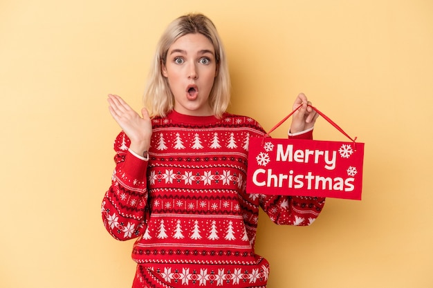 Junge kaukasische Frau, die ein Plakat der frohen Weihnachten lokalisiert auf gelbem Hintergrund hält überrascht und schockiert.