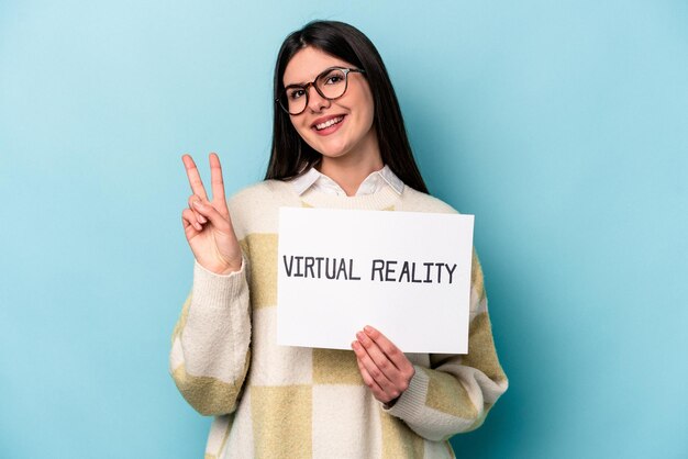 Junge kaukasische Frau, die ein Plakat der virtuellen Realität hält, das auf blauem Hintergrund isoliert ist, fröhlich und sorglos, zeigt ein Friedenssymbol mit den Fingern