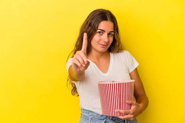 Junge kaukasische Frau, die ein Popcorn hält, das auf gelbem Hintergrund isoliert ist und Nummer eins mit dem Finger zeigt.