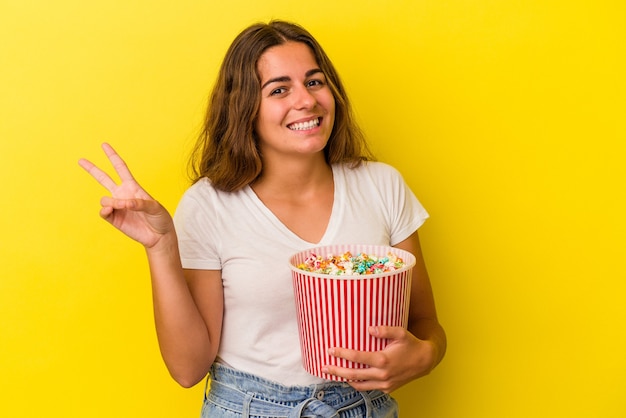 Junge kaukasische Frau, die ein Popcorn isoliert auf gelbem Hintergrund hält, fröhlich und sorglos, das ein Friedenssymbol mit den Fingern zeigt.