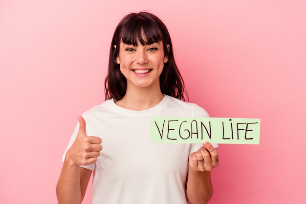 Foto junge kaukasische frau, die ein veganes lebensplakat lokalisiert, das auf rosa hintergrund lächelt und daumen oben anhebt