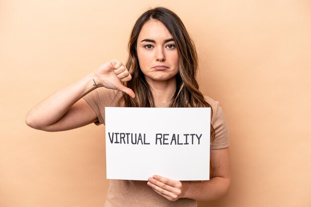 Junge kaukasische Frau, die ein Virtual-Reality-Plakat hält, das auf beigem Hintergrund isoliert ist und eine Abneigungsgeste mit dem Daumen nach unten zeigt Meinungsverschiedenheitskonzept