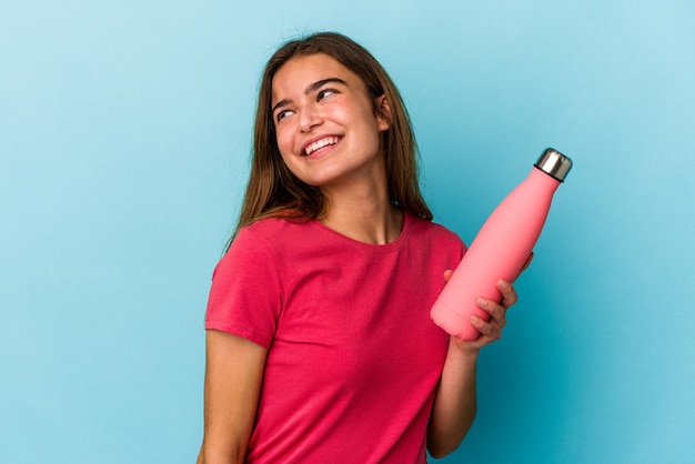 Junge kaukasische Frau, die eine auf blauem Hintergrund isolierte Wasserflasche hält, sieht beiseite lächelnd, fröhlich und angenehm aus.