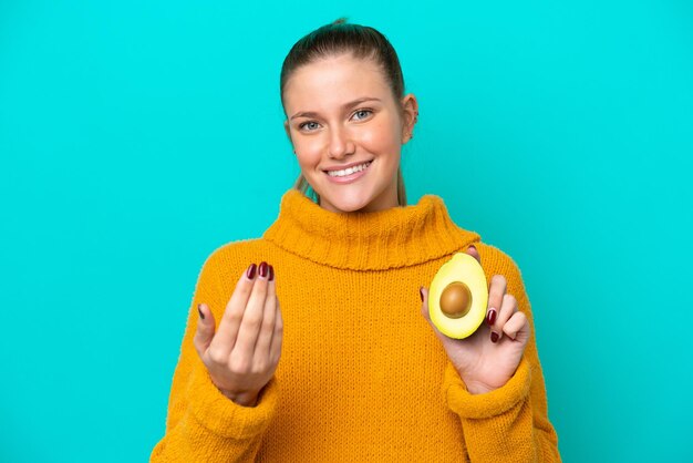 Junge kaukasische Frau, die eine Avocado isoliert auf blauem Hintergrund hält und einlädt, mit der Hand zu kommen Glücklich, dass Sie gekommen sind