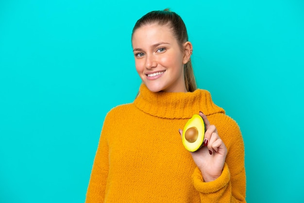 Junge kaukasische Frau, die eine Avocado isoliert auf blauem Hintergrund hält und viel lächelt