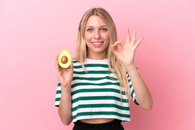 Junge kaukasische Frau, die eine Avocado isoliert auf rosafarbenem Hintergrund hält und mit den Fingern ein Ok-Zeichen zeigt