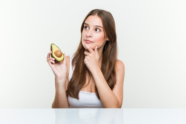 Junge kaukasische Frau, die eine Avocado seitlich schaut mit zweifelhaftem und skeptischem Ausdruck anhält.