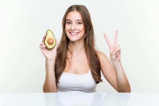 Junge kaukasische Frau, die eine Avocado zeigt Nummer zwei mit den Fingern hält.