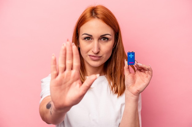 Junge kaukasische Frau, die eine Batterie isoliert auf rosafarbenem Hintergrund hält, die mit ausgestreckter Hand steht und ein Stoppschild zeigt, was Sie daran hindert.