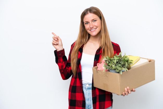 Junge kaukasische Frau, die eine Bewegung macht, während sie eine Kiste voller Dinge aufhebt, die auf weißem Hintergrund isoliert sind und mit dem Finger zur Seite zeigt