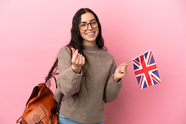 Junge kaukasische Frau, die eine Flagge des Vereinigten Königreichs isoliert auf rosa Hintergrund hält und Geldgeste macht