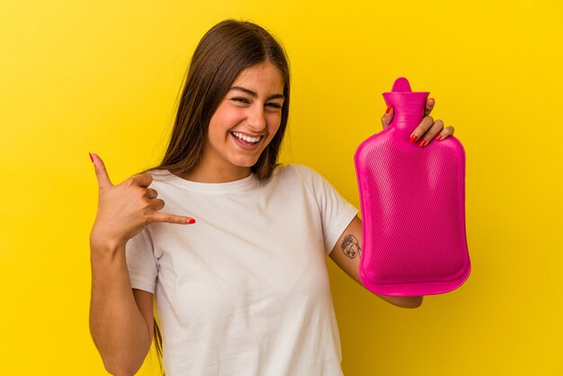 Junge kaukasische Frau, die eine heiße Flasche Wasser hält, die auf gelbem Hintergrund lokalisiert wird und eine Handyanrufgeste mit den Fingern zeigt.