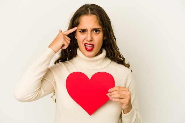 Junge kaukasische Frau, die eine Herz-Valentinstagform lokalisiert hält, die eine Enttäuschungsgeste mit Zeigefinger zeigt.