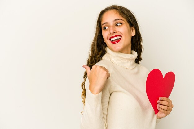 Junge kaukasische Frau, die eine Herz-Valentinstagform lokalisierte Punkte mit Daumenfinger weg, lachend und sorglos hält.