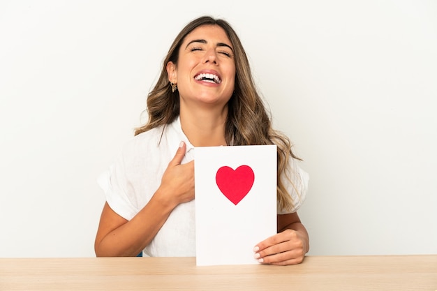 Junge kaukasische Frau, die eine isolierte Valentinstagskarte hält, lacht laut und hält Hand auf Brust.