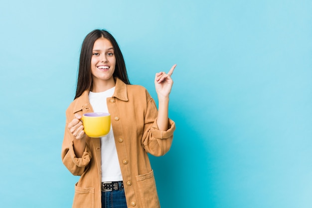 Junge kaukasische Frau, die eine Kaffeetasse hält, die fröhlich mit Zeigefinger weg zeigt.