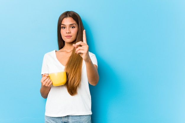 Junge kaukasische Frau, die eine Kaffeetasse hält, die Nummer eins mit Finger zeigt.