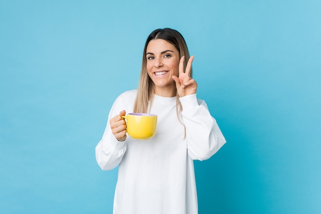 Junge kaukasische Frau, die eine Kaffeetasse zeigt Siegeszeichen anhält und breit lächelt.