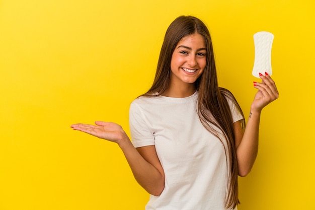 Junge kaukasische Frau, die eine Kompresse isoliert auf gelber Wand hält, die einen Kopienraum auf einer Handfläche zeigt und eine andere Hand an der Taille hält.