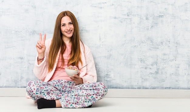 Junge kaukasische Frau, die eine Müslischale hält, die Nummer zwei mit den Fingern zeigt.