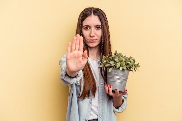 Foto junge kaukasische frau, die eine pflanze isoliert auf gelbem hintergrund hält, die mit ausgestreckter hand steht und ein stoppschild zeigt, das sie verhindert