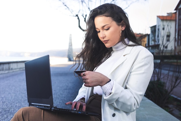 Junge kaukasische Frau, die eine rote Kreditkarte verwendet, um online mit einem Laptop auf der Straße zu bezahlen