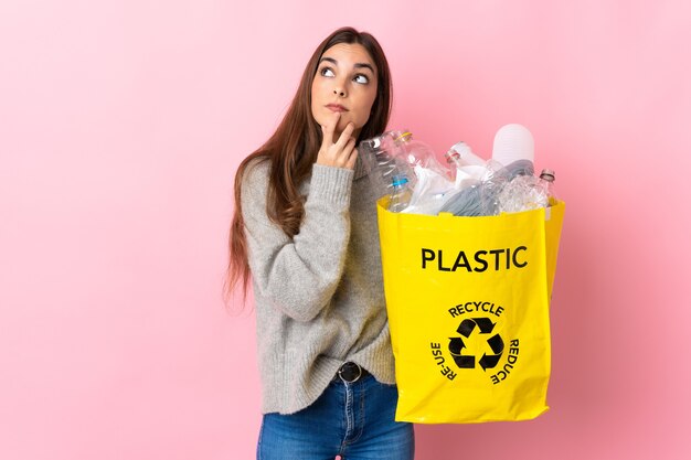 Junge kaukasische Frau, die eine Tasche voll von Plastikflaschen hält, um lokalisiert auf Rosa zu recyceln, das Zweifel beim Aufblicken hat