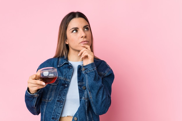 Junge kaukasische Frau, die eine Teetasse hält, die seitwärts mit zweifelhaftem und skeptischem Ausdruck schaut.