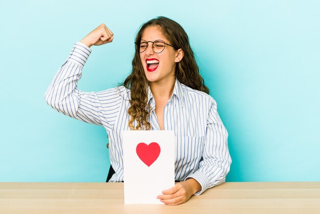 Junge kaukasische Frau, die eine Valentinstagskarte hält, die erhöhte Faust nach einem Sieg, Gewinnerkonzept isoliert.