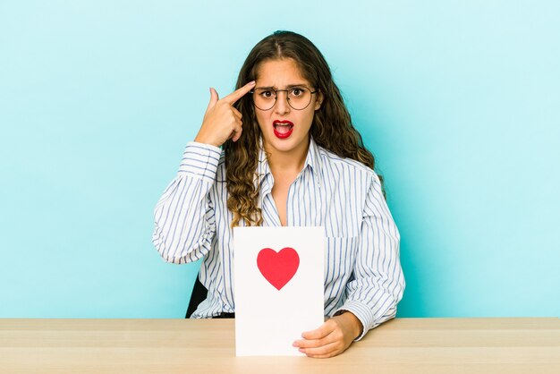 Junge kaukasische Frau, die eine Valentinstagskarte lokalisiert hält, die eine Enttäuschungsgeste mit Zeigefinger zeigt.