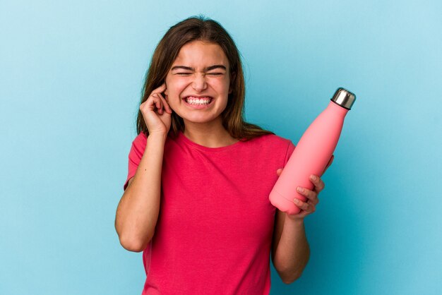 Junge kaukasische Frau, die eine Wasserflasche lokalisiert auf blauem Hintergrund hält, der Ohren mit den Händen bedeckt.