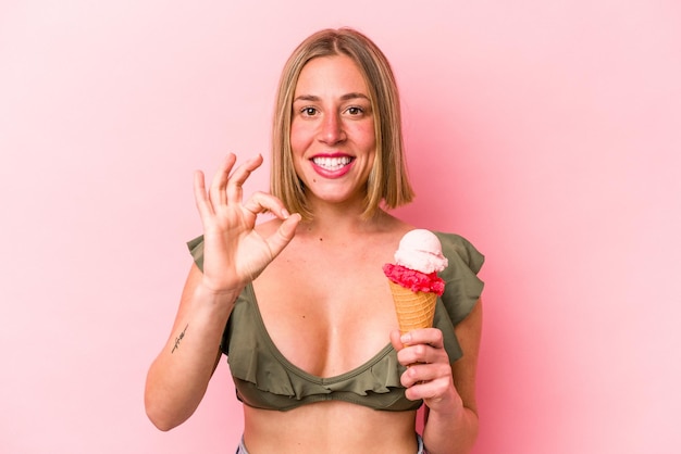Junge kaukasische Frau, die einen Bikini trägt und ein Eis hält, das auf rosa Hintergrund isoliert ist, fröhlich und selbstbewusst und zeigt eine ok-Geste