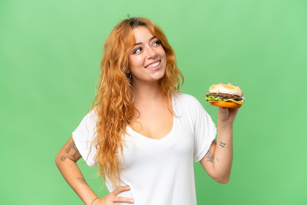 Junge kaukasische Frau, die einen Burger isoliert auf grünem Chroma-Key-Hintergrund hält und lächelnd nach oben schaut