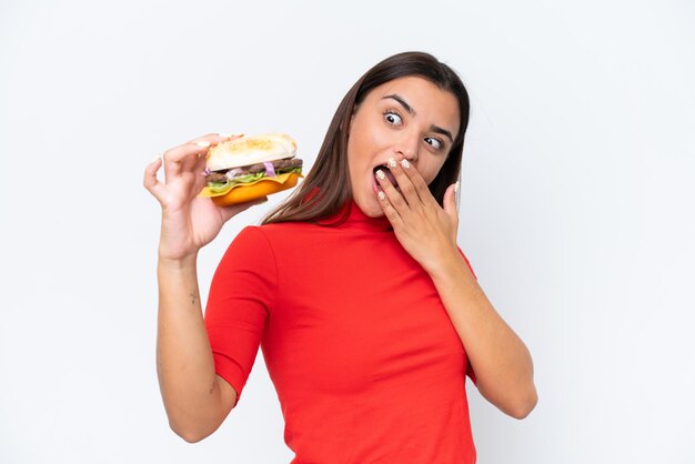 Junge kaukasische Frau, die einen Burger isoliert auf weißem Hintergrund mit überraschtem und schockiertem Gesichtsausdruck hält