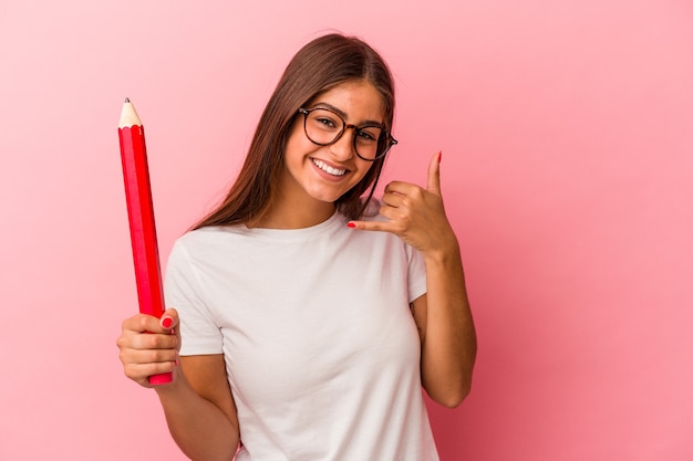 Junge kaukasische Frau, die einen großen Bleistift hält, der auf rosafarbenem Hintergrund lokalisiert wird und eine Handyanrufgeste mit den Fingern zeigt.