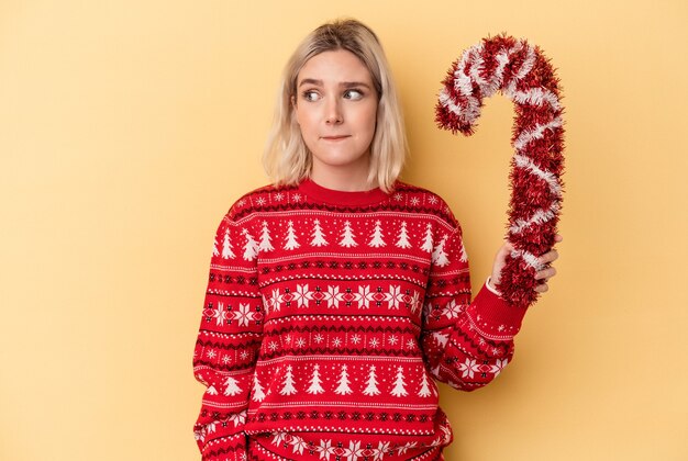 Junge kaukasische Frau, die einen großen Weihnachtsstab einzeln auf gelbem Hintergrund hält, verwirrt, fühlt sich zweifelhaft und unsicher.