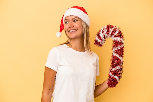 Junge kaukasische Frau, die einen großen Weihnachtsstab isoliert auf gelbem Hintergrund hält, sieht beiseite lächelnd, fröhlich und angenehm aus.