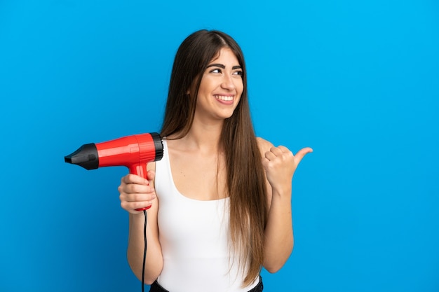 Junge kaukasische Frau, die einen Haartrockner isoliert auf blauem Hintergrund hält und auf die Seite zeigt, um ein Produkt zu präsentieren?