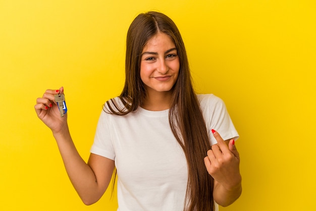 Junge kaukasische Frau, die einen Home-Schlüssel einzeln auf gelbem Hintergrund hält und mit dem Finger auf Sie zeigt, als ob sie einladen würde, näher zu kommen.