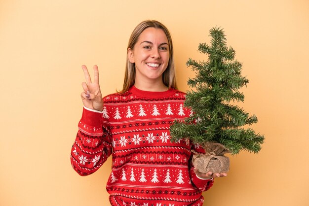 Junge kaukasische Frau, die einen kleinen Weihnachtsbaum auf gelbem Hintergrund hält, der Nummer zwei mit den Fingern zeigt.