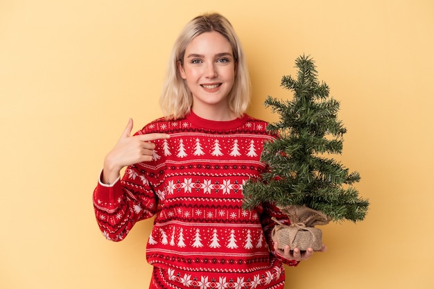 Junge kaukasische Frau, die einen kleinen Weihnachtsbaum isoliert auf gelbem Hintergrund hält, der mit der Hand auf einen Hemdkopierraum zeigt, stolz und selbstbewusst