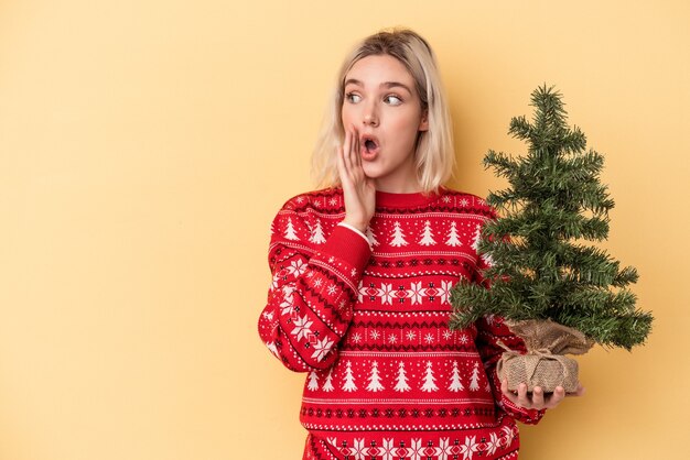 Junge kaukasische Frau, die einen kleinen Weihnachtsbaum isoliert auf gelbem Hintergrund hält, sagt eine geheime heiße Bremsnachricht und schaut beiseite
