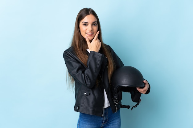 Junge kaukasische Frau, die einen Motorradhelm auf Blau glücklich und lächelnd hält