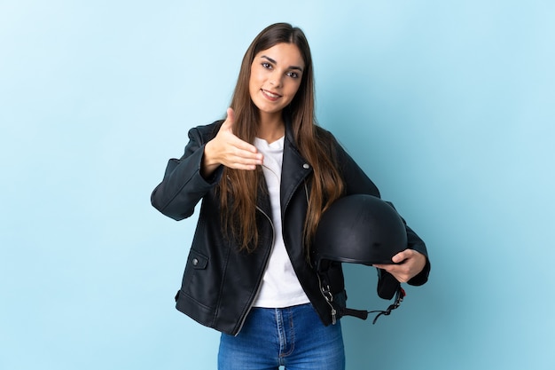 Junge kaukasische Frau, die einen Motorradhelm lokalisiert auf blauen Händeschütteln für das Schließen eines guten Geschäfts hält