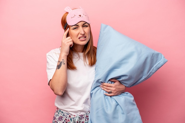 Junge kaukasische Frau, die einen Pyjama trägt und Kissen isoliert auf rosa Hintergrund hält und eine Enttäuschungsgeste mit dem Zeigefinger zeigt.