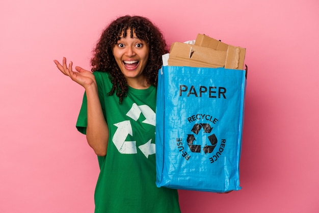 Junge kaukasische Frau, die einen recycelten Kunststoff lokalisiert auf rosa Wand hält, die eine angenehme Überraschung empfängt, aufgeregt und Hände hebt.