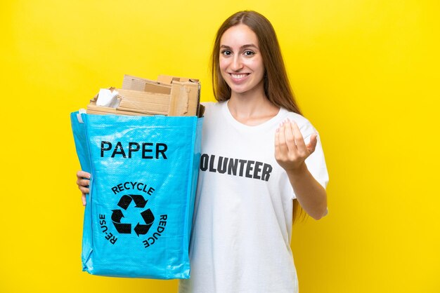 Junge kaukasische Frau, die einen Recycling-Beutel voller Papier hält, um isoliert auf gelbem Hintergrund zu recyceln, der einlädt, mit der Hand zu kommen Glücklich, dass Sie gekommen sind